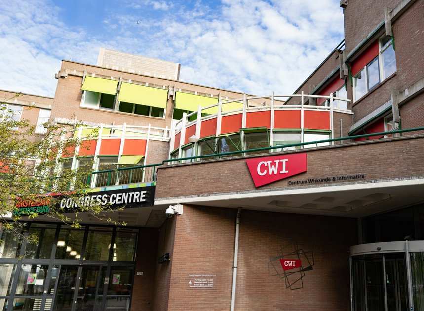 A photograph of the CWI building, the national research institute for mathematics and computer science in the Netherlands. The building has multiple levels with a combination of green, orange, and red panels around the windows, creating a colorful façade. The CWI logo is prominently displayed above the entrance and on a sign projecting from the building. Below, the Amsterdam Congress Centre sign stretches across the front over the glass doors. The sky above the building is clear and blue with a few clouds, hinting at a pleasant day.                            